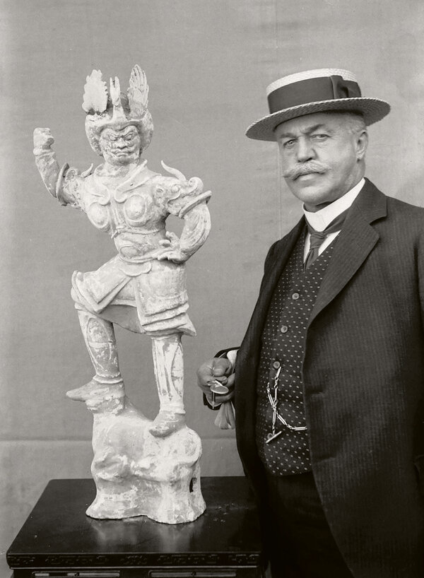 Foto von Adolf Fischer, mit Hut neben einer Statue stehend.