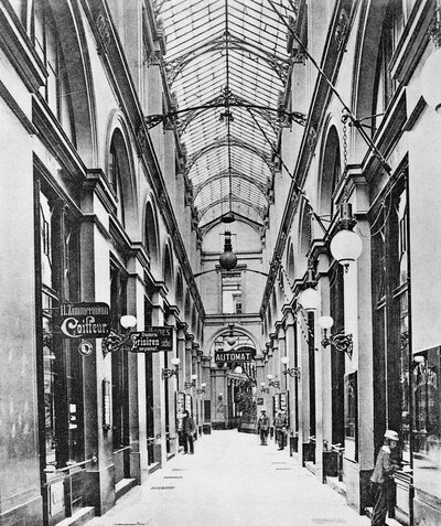 Historische Innenansicht einer lang gezogenen Einkaufspassage mit hohem Glasdach, filigraner Metallkonstruktion und Ladenfronten auf beiden Seiten.