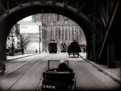 Schwarz-Weiß-Filmszene mit Blick durch einen dunklen Torbogen auf eine Straße. Im Vordergrund steht ein historisches Automobil, dahinter sind Gebäude zu erkennen.