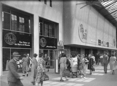 Historische Schwarz-Weiß-Aufnahme des Aktualitätenkinos im Kölner Hauptbahnhof. Menschen stehen und gehen vor dem Kinoeingang, an dessen Fassade Filmplakate hängen.