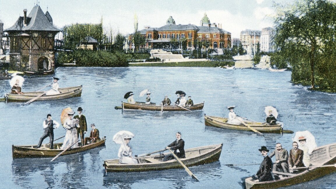 Historische Fotografie zeigt Personen in mehreren Ruderboote auf einem See. Im Hintergrund befinden sich Gebäude.