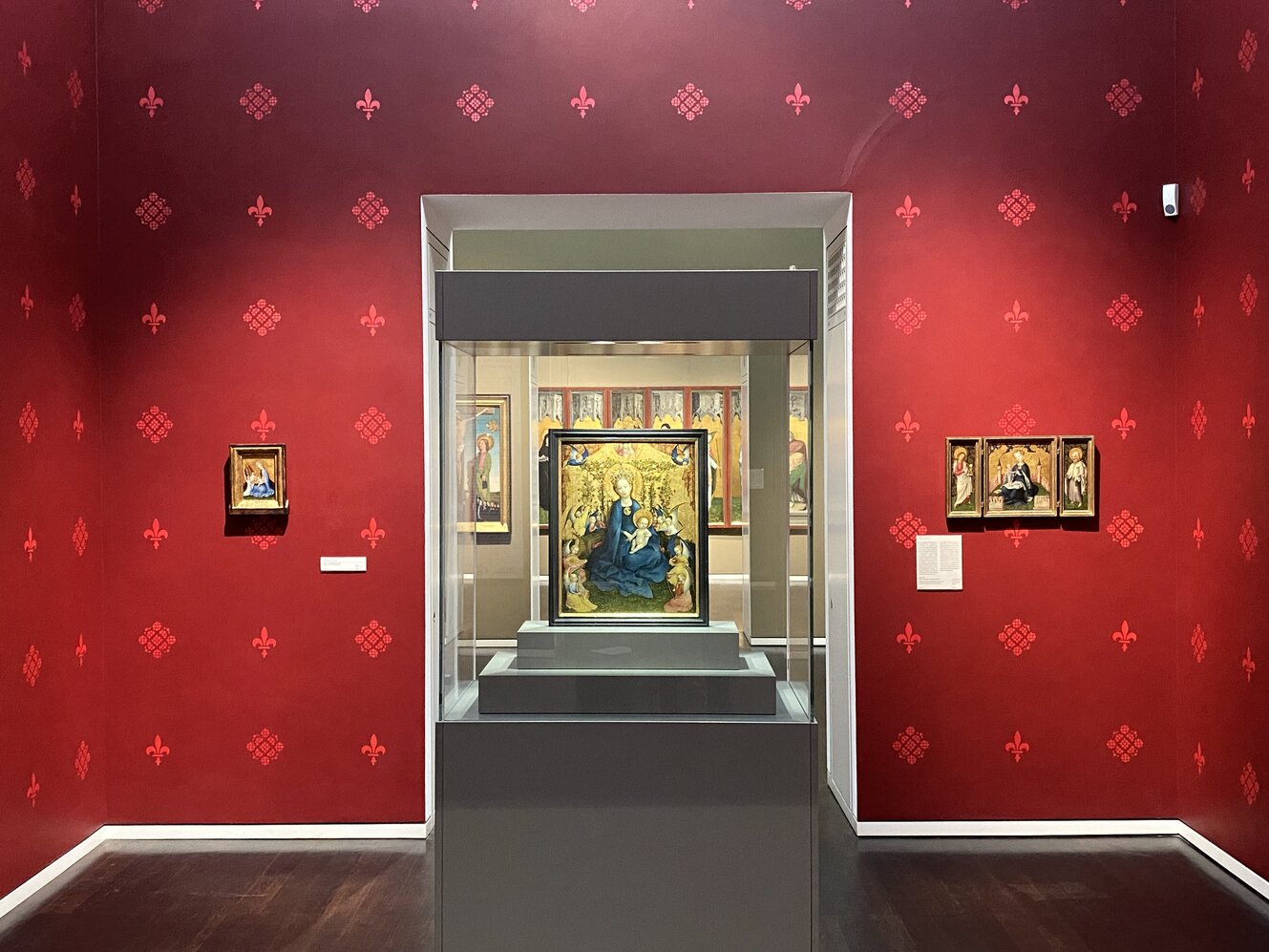 Blick in die Ausstellungsräume. In der Mitte steht eine Vitrine mit Lochners Madonna im Rosenhag. Die Wände sind rot tapeziert und gemustert. 