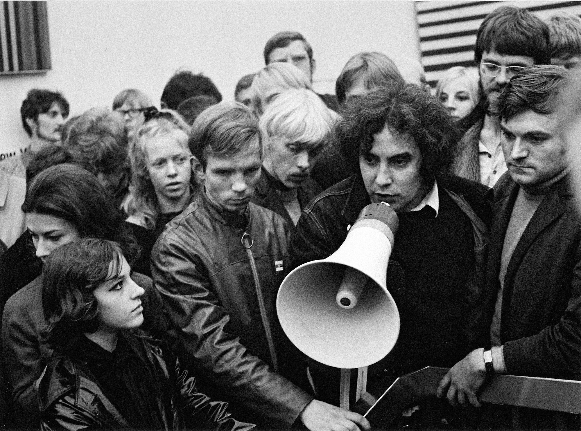 Auf dem schwarz-weiß Foto ist eine Menschenmenge abgebildet. In der Mitte steht ein Mann mit Megafon. 